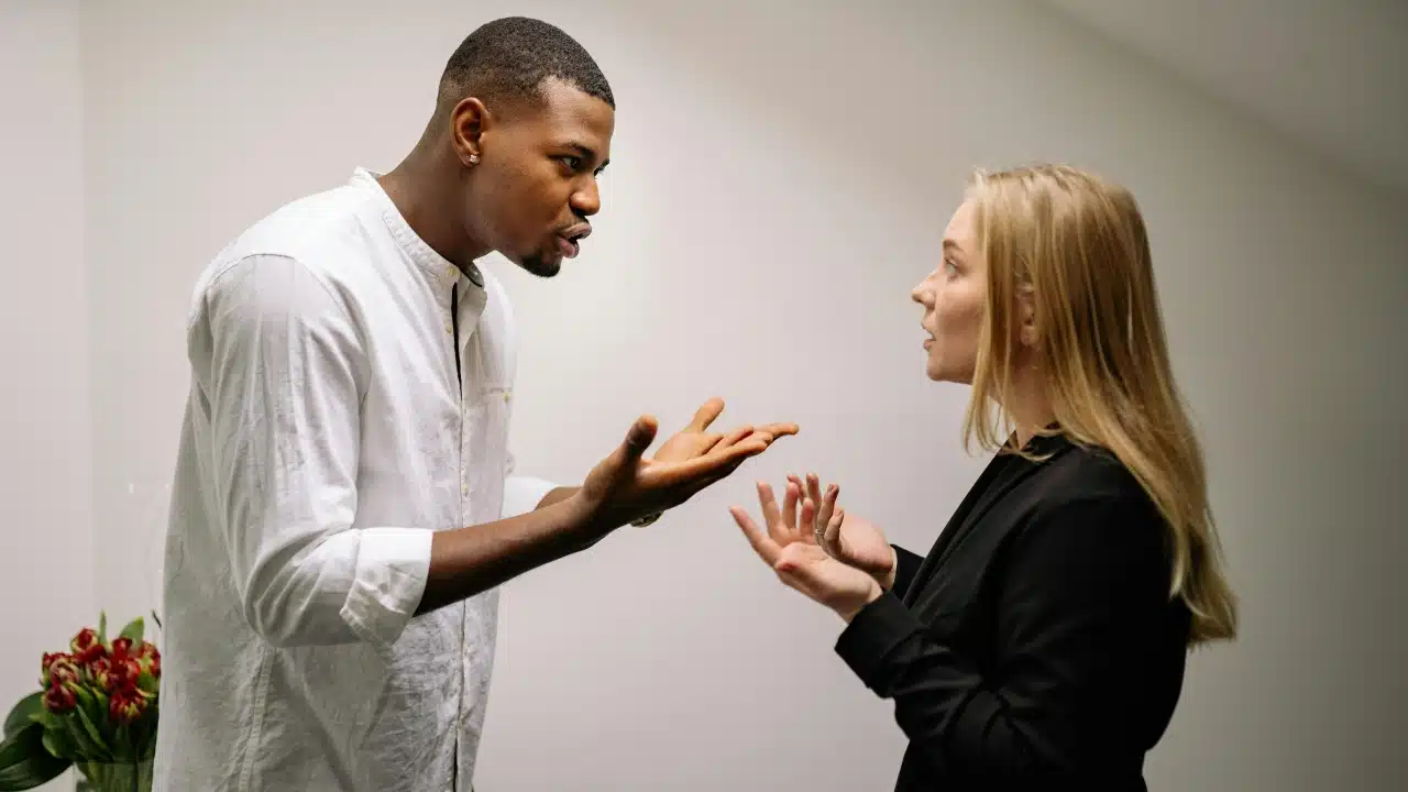A man and woman stand facing each other while gesturing with open palms during a tense conversation.