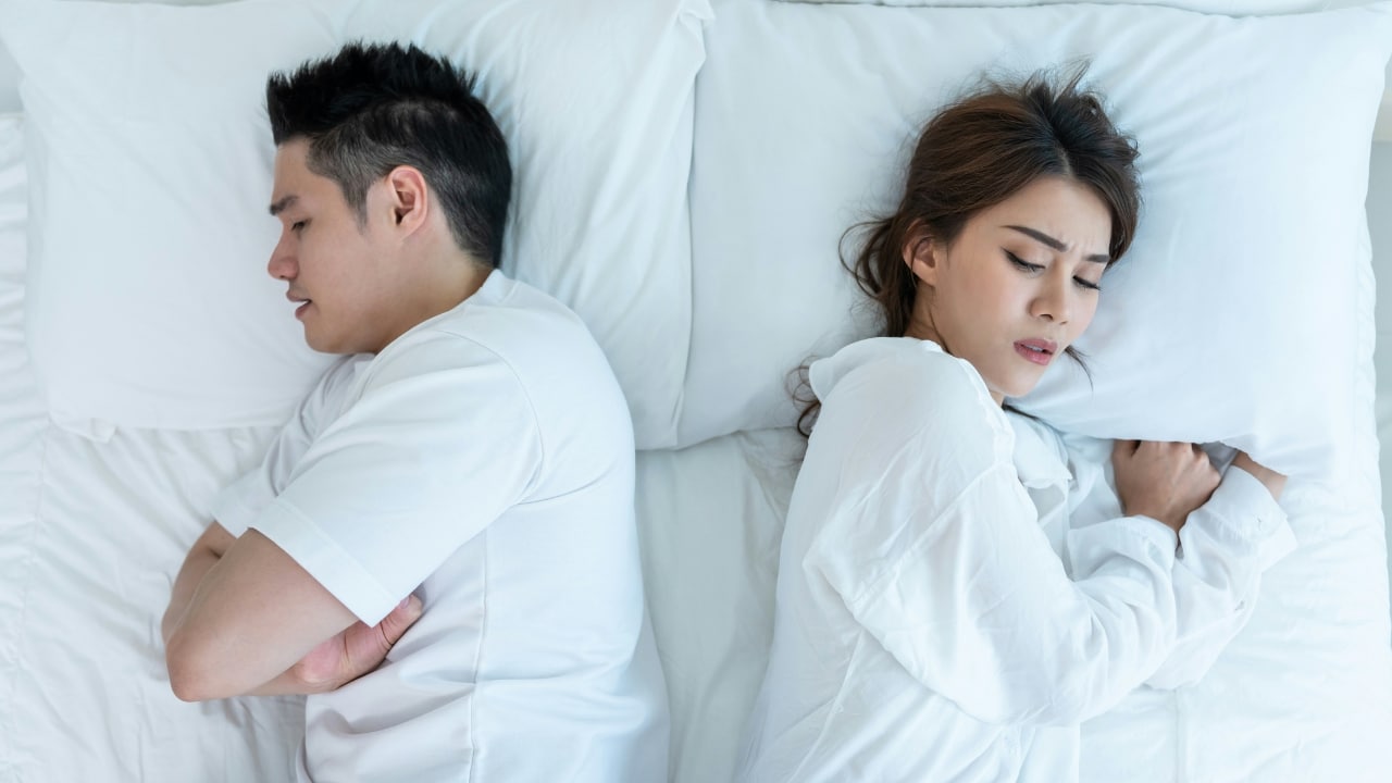A man and woman lie in bed facing away from each other with distressed expressions.