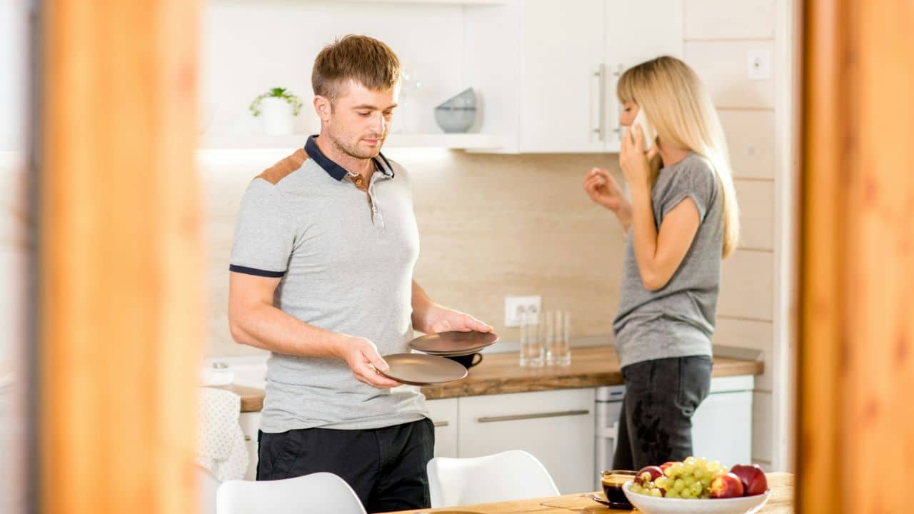 A man carries plates in a kitchen while a woman talks on a cellphone.