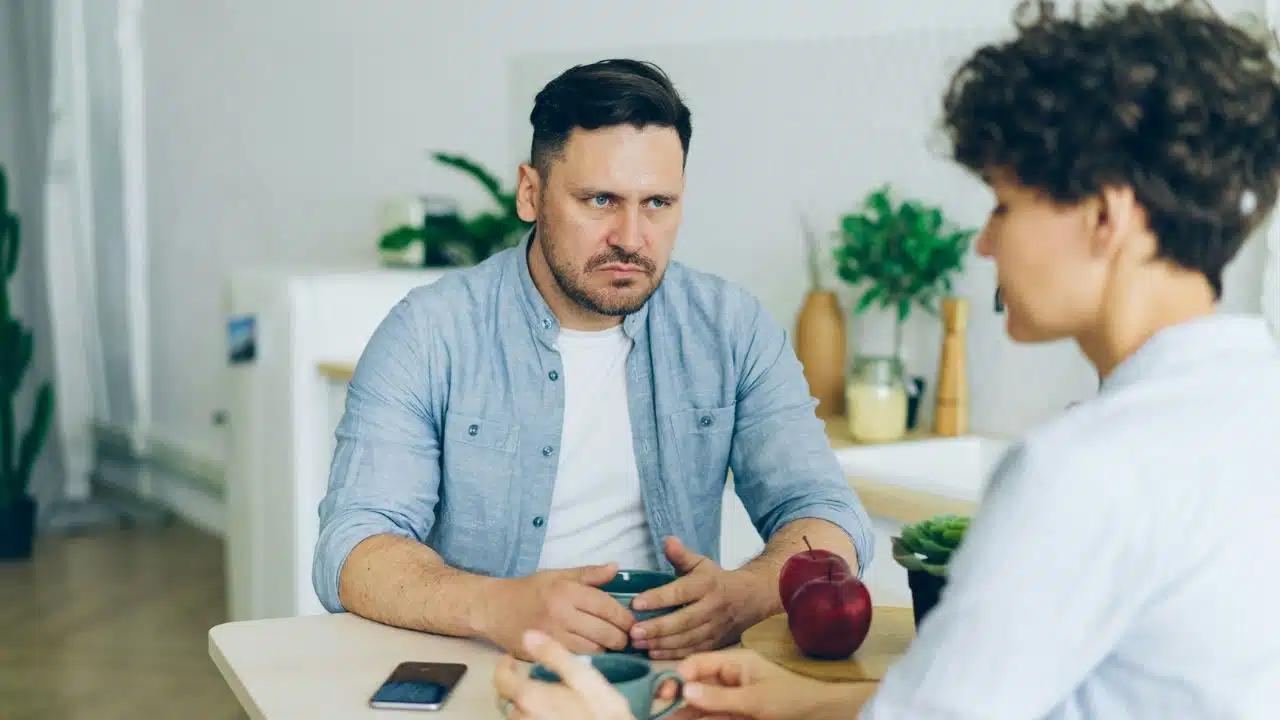 A man with a serious expression sits at a table across from a woman holding a cup.