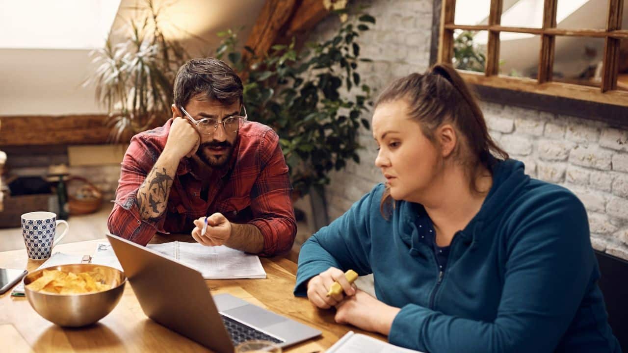 Two people sit at a table looking at a laptop screen next to a bowl.