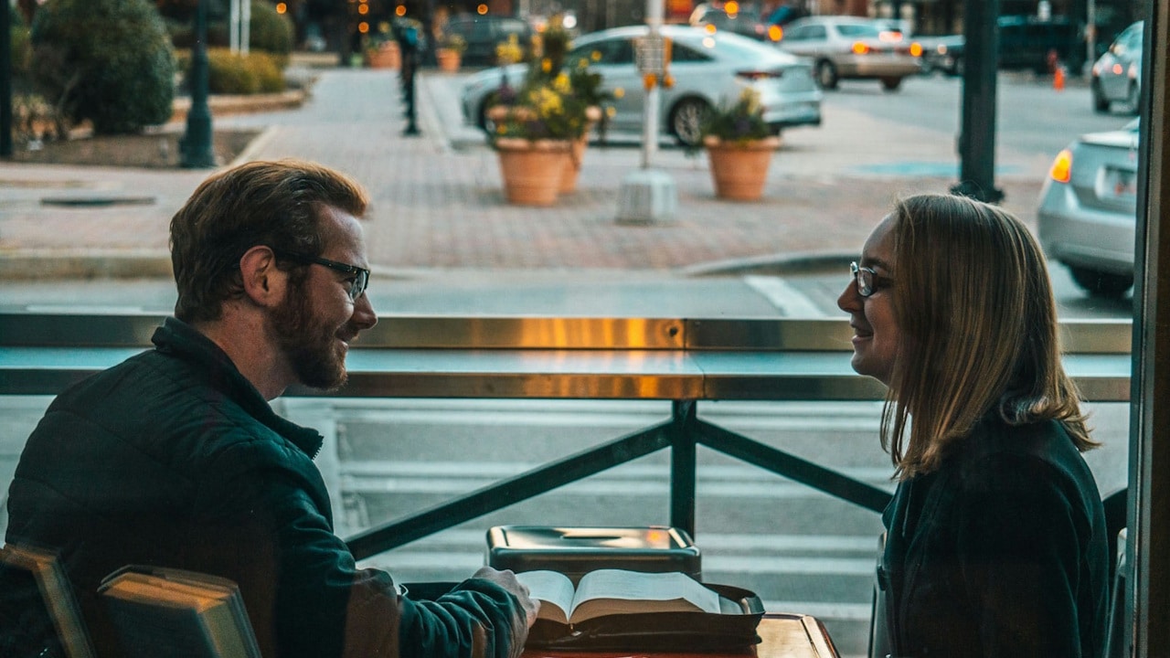 A man and a woman wearing glasses smiling at each other across a table by a window.