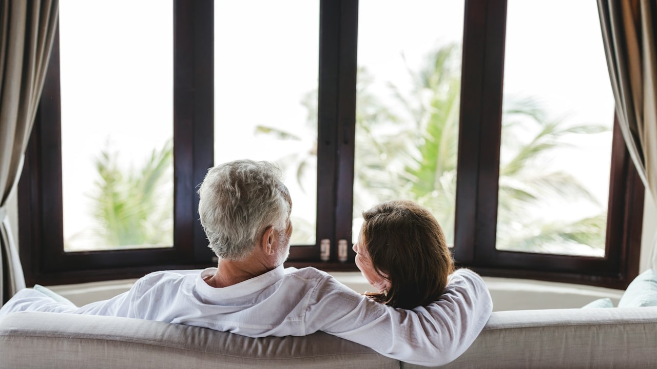 An older couple sitting on a couch, seen from behind, looking out a large window at palm trees and bright light.