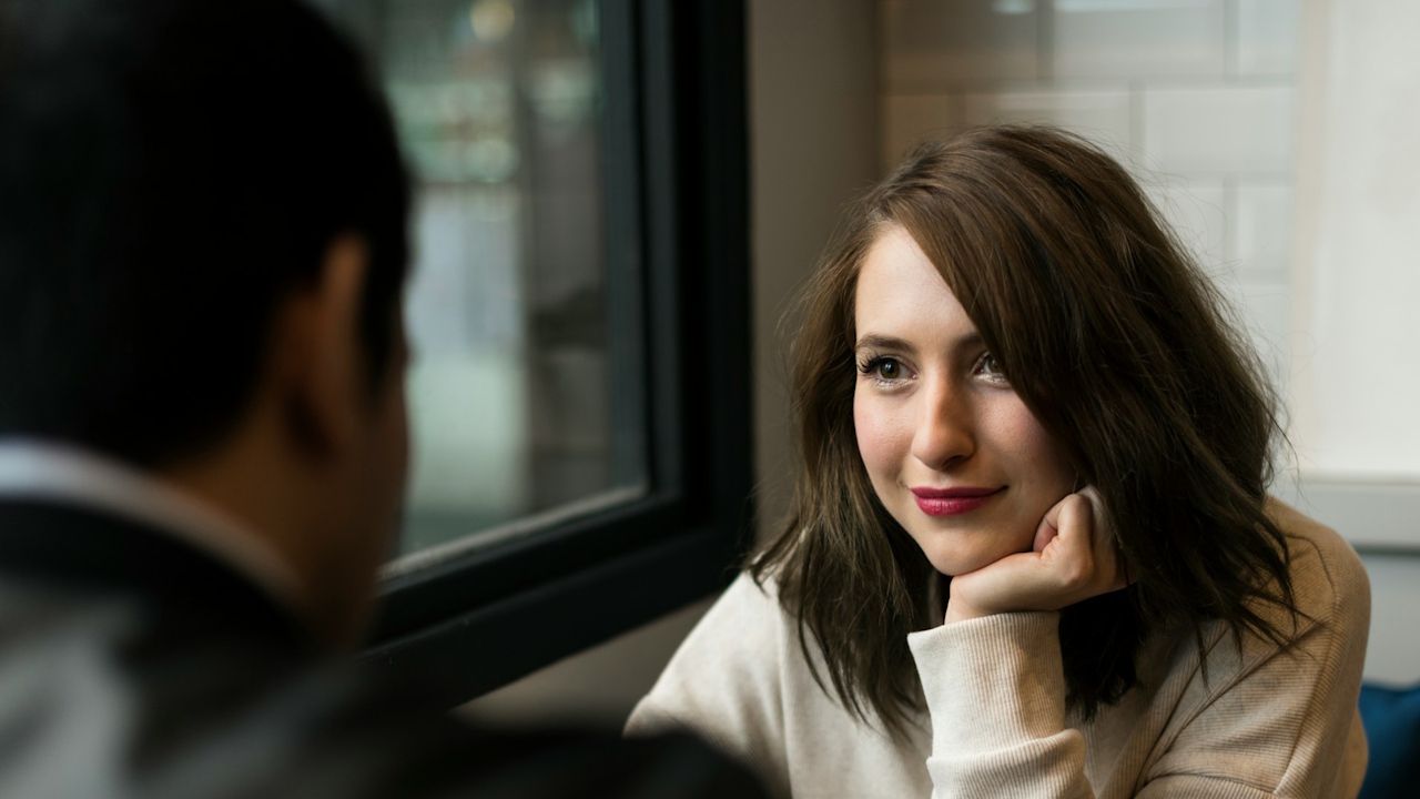 A woman smiling and resting her chin on her hand while talking to a man in a coffee shop.