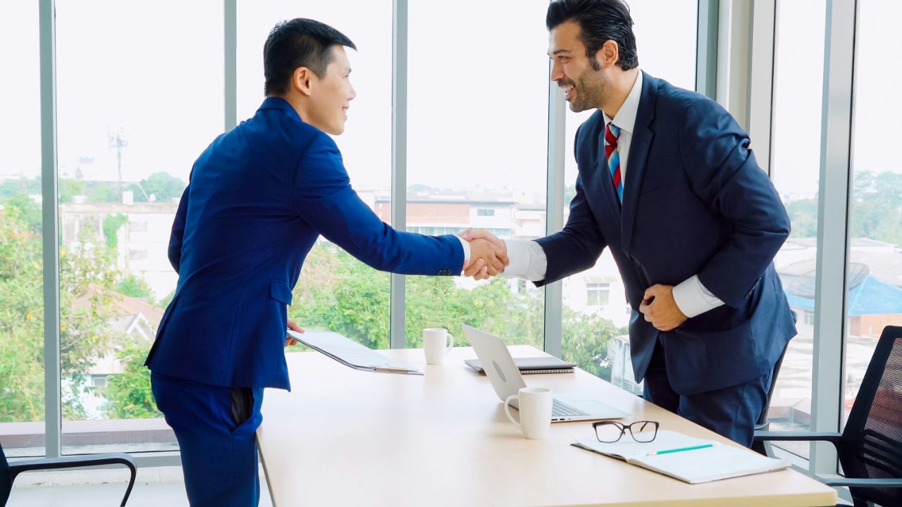 Two men in business suits are shaking hands across a long desk in a bright office.