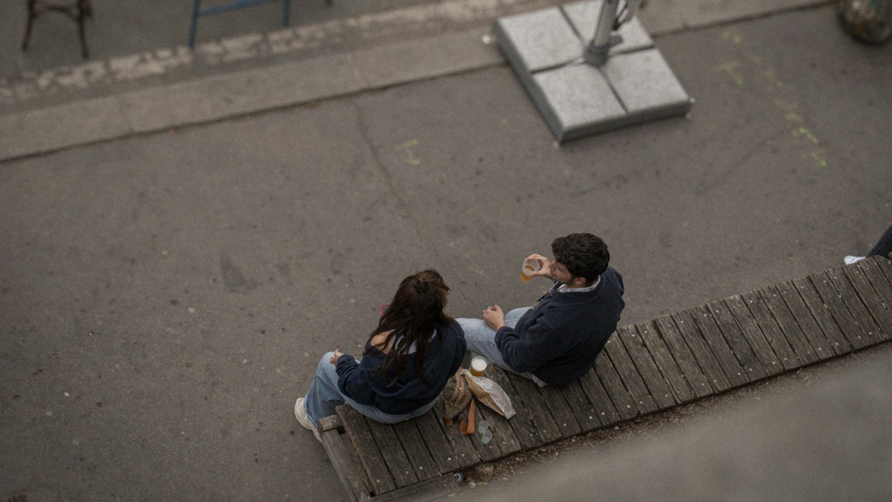 Two people sitting on a wooden platform near a street, viewed from above.