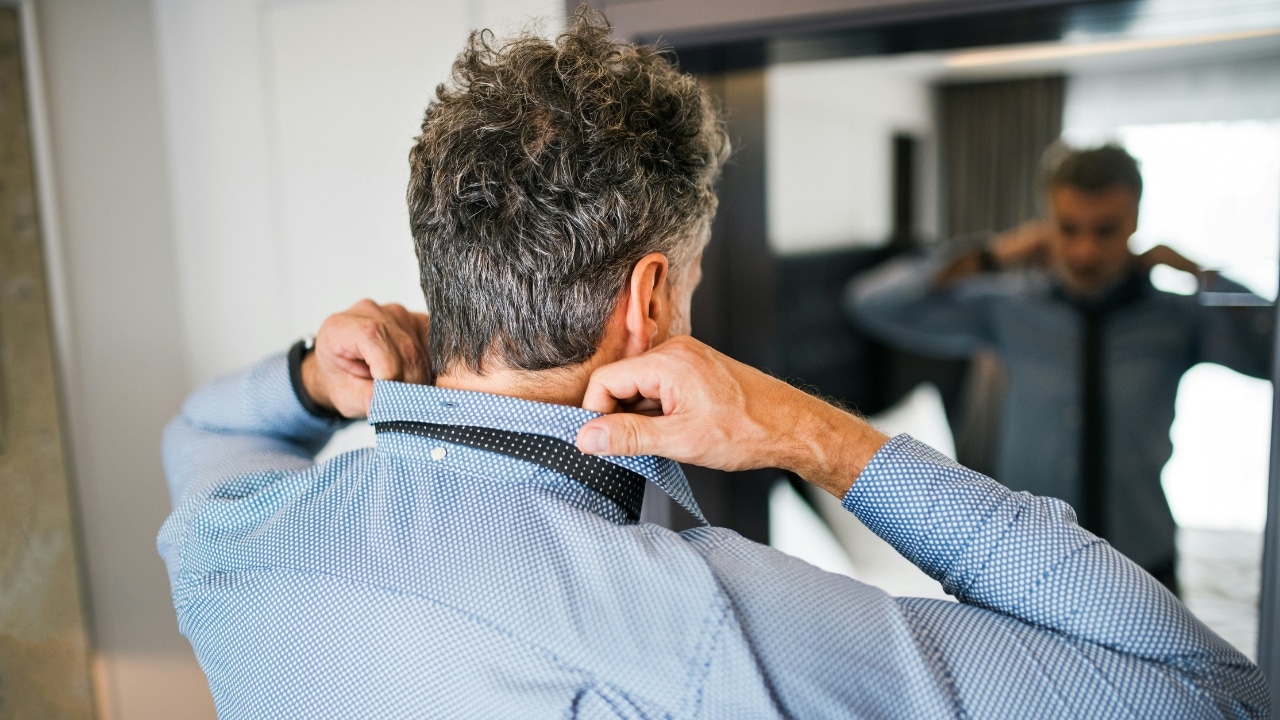 A man stands before a mirror while adjusting the collar of his blue patterned shirt.