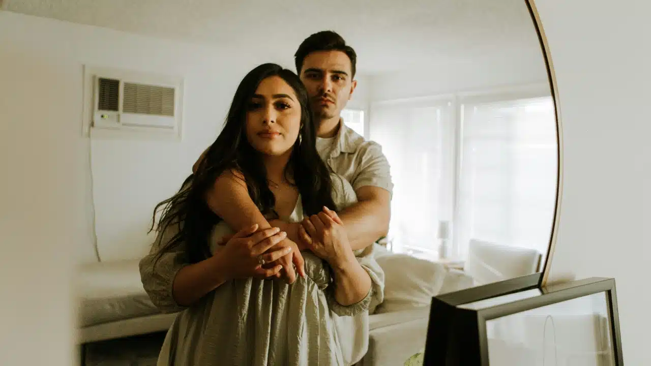 A man stands behind a woman and embraces her while they look into a mirror.
