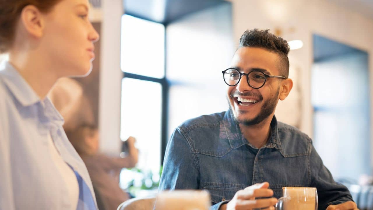 A man with glasses laughs while sitting at a table across from a woman.
