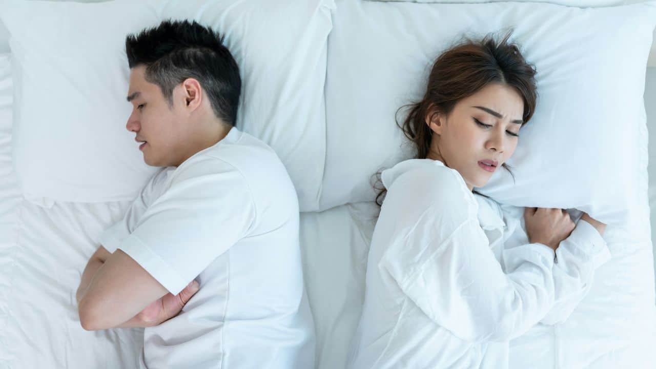 A man and woman lie in bed facing away from each other with eyes closed.