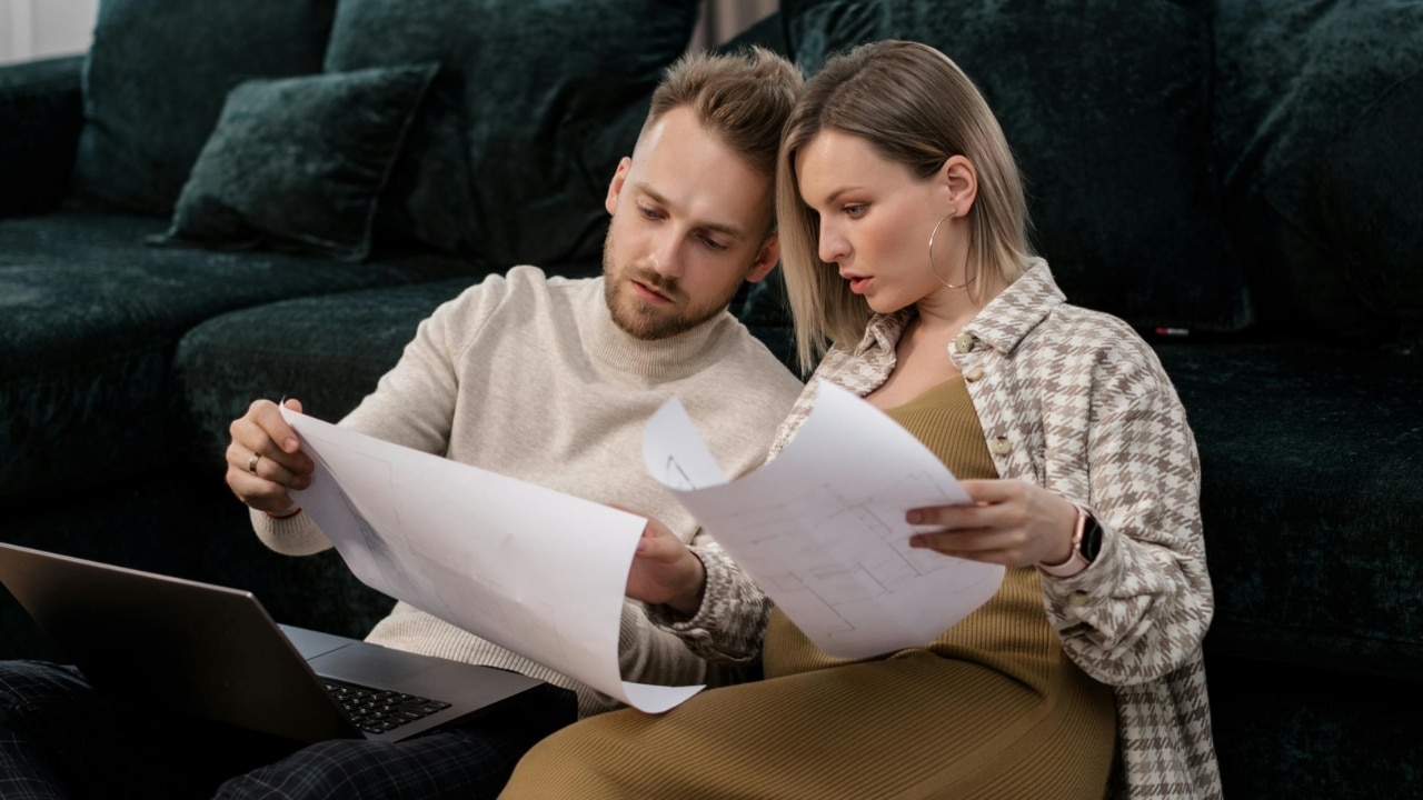 A man and woman sit on the floor looking at large papers next to a laptop.