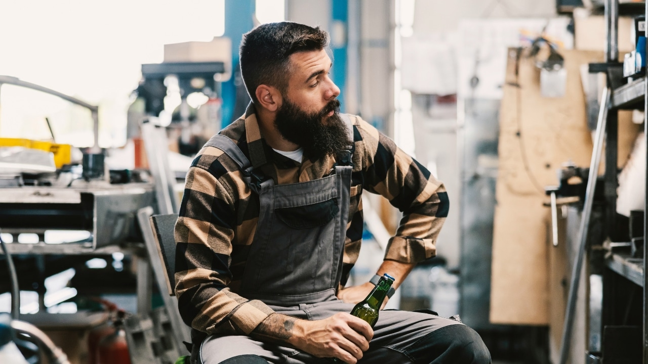 A bearded man in overalls sits in a workshop holding a green glass bottle of beer.