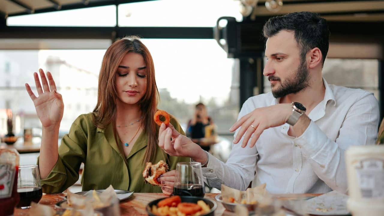 A woman holding a burger looks down while a man offers her an onion ring.