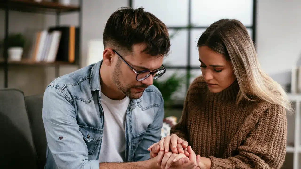 A man and woman sit close together with their heads bowed and hands clasped together.