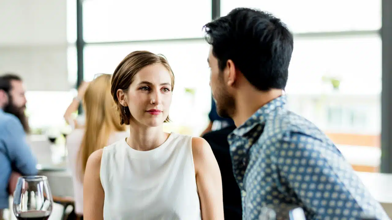 A woman in a white top looks toward a man in a blue patterned shirt.