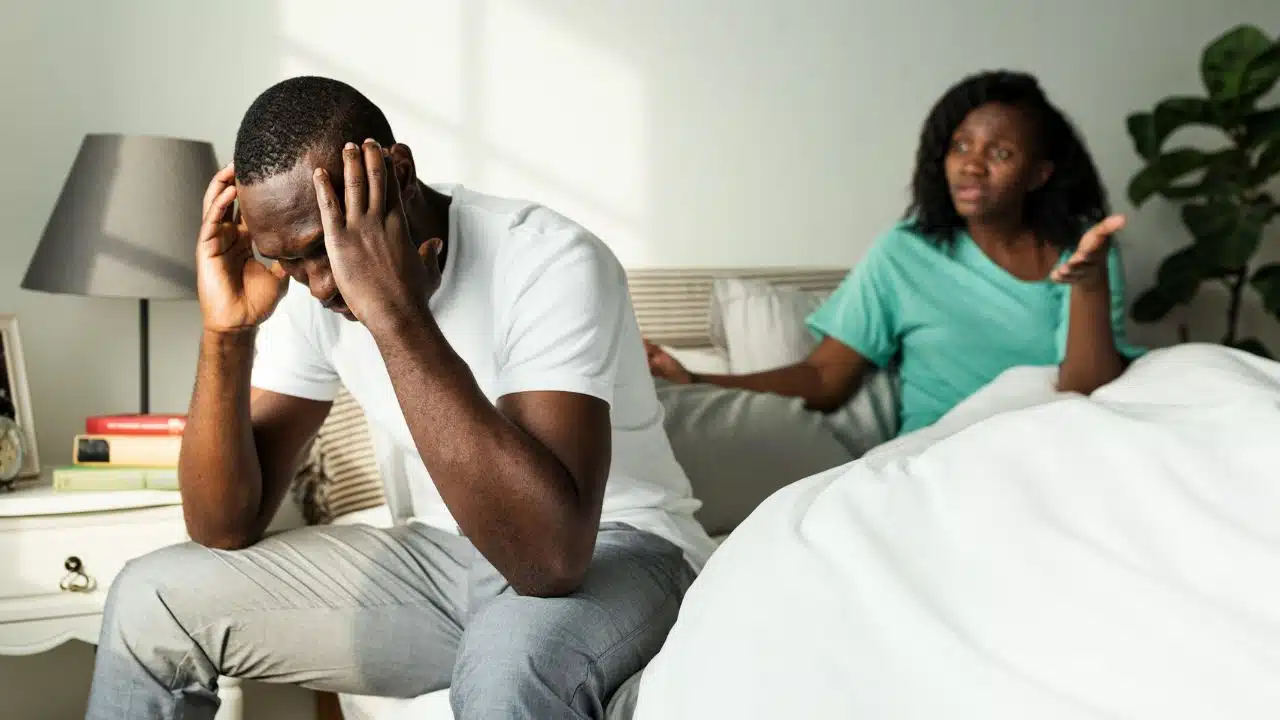 A man sits on a bed with his head in his hands while a woman gestures.