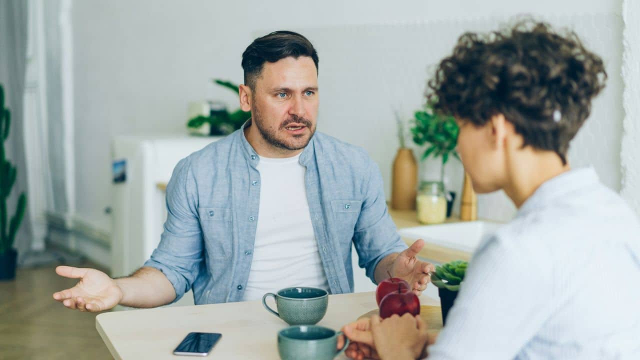 A man gestures with open hands while speaking to a woman at a kitchen table.