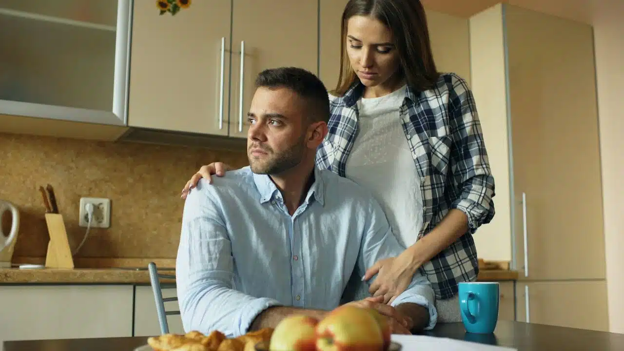 A woman stands behind a seated man in a kitchen with her hand on his shoulder