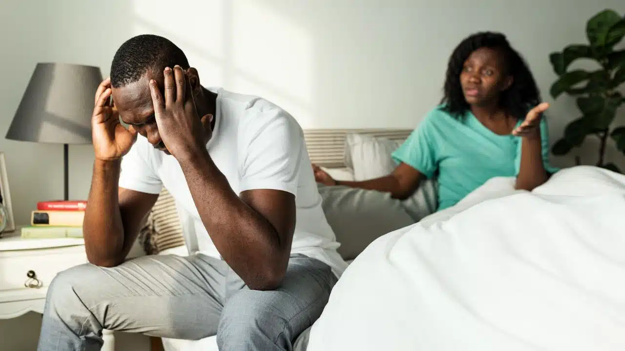 A man sits on a bed holding his head while a woman gestures behind him.