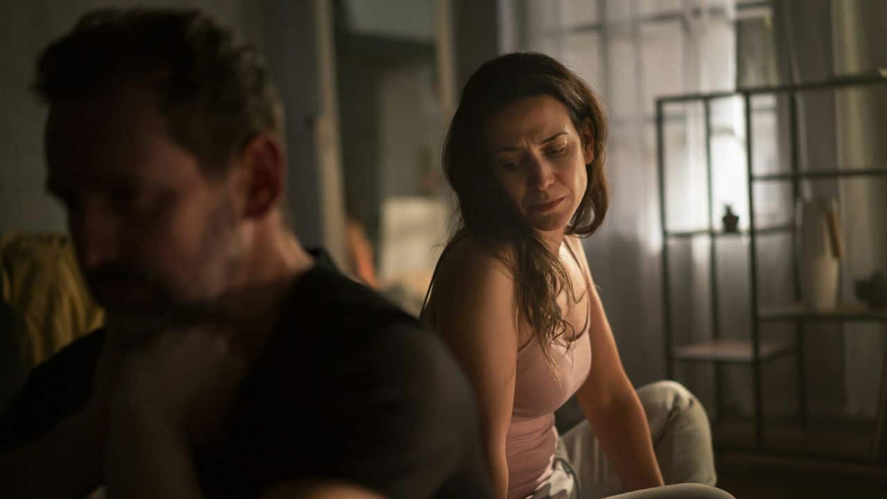 A woman looks down sadly toward a man sitting in the foreground with his back turned.