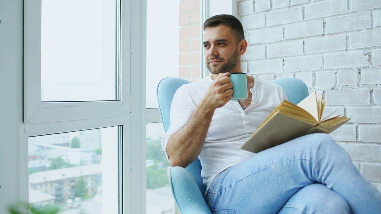 A man sits in a blue chair by a window, holding a mug and book.