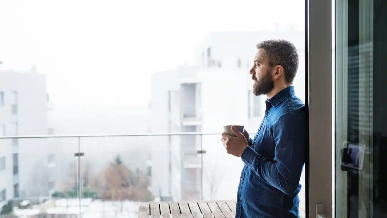 A bearded man in a blue shirt holds a mug while looking out from a balcony.