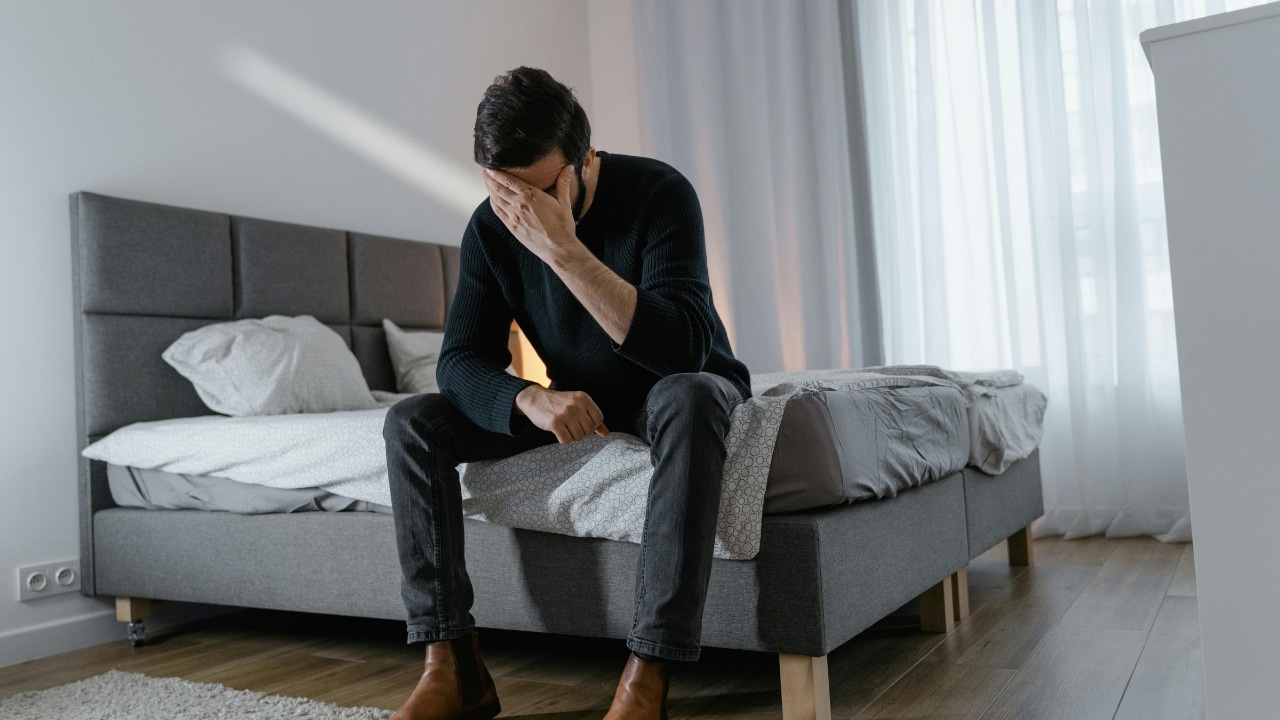 A man in a dark sweater sits on the edge of a bed with his head down.