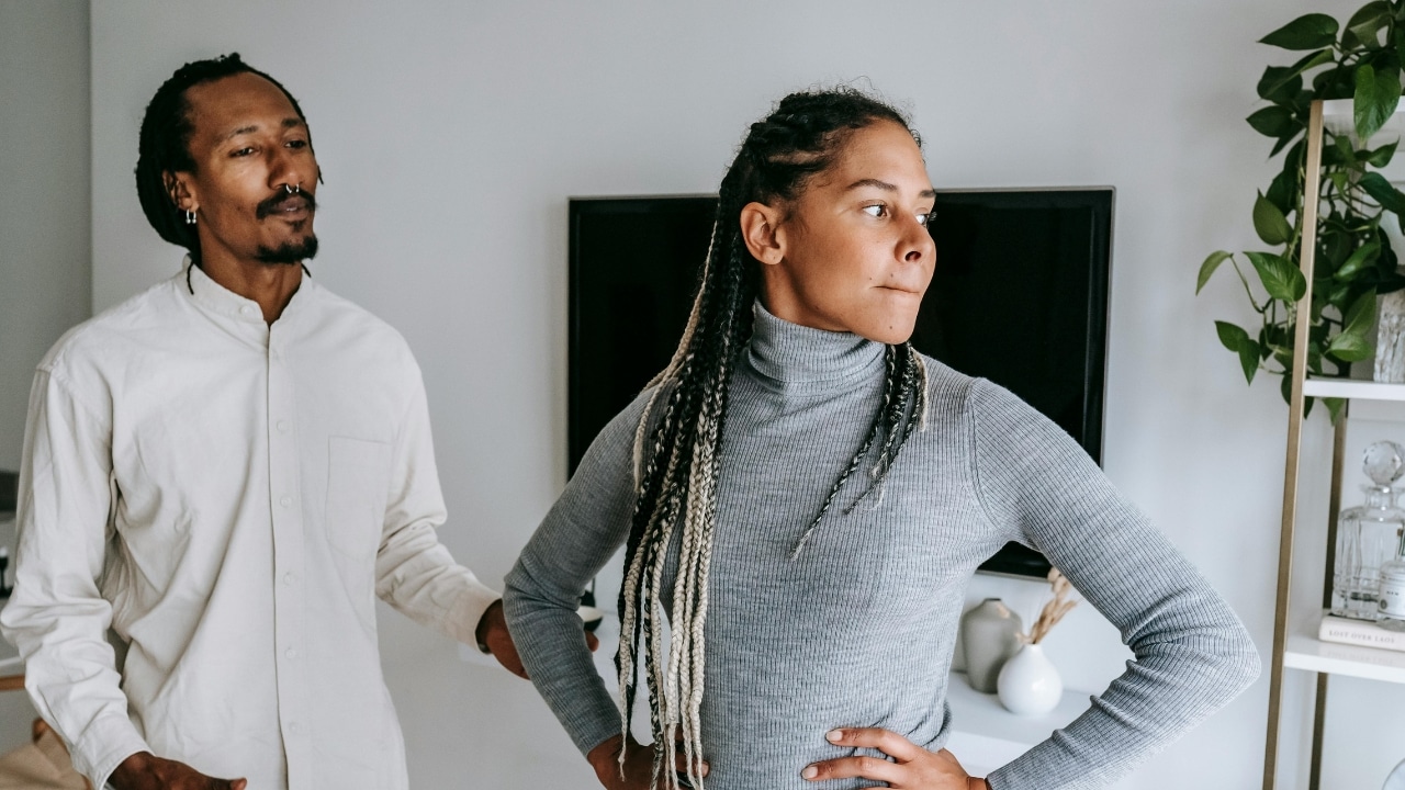 A woman with long braids looks away while a man stands behind her in a room.