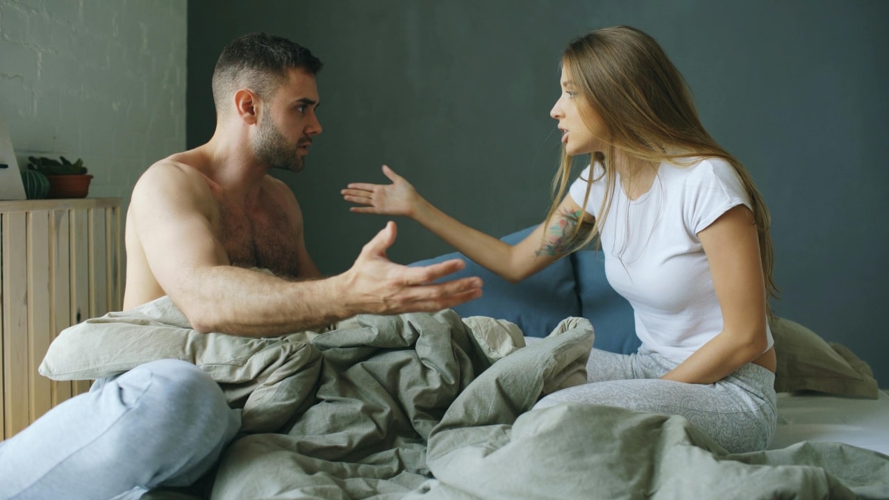 A man and woman sit on a bed, gesturing with their hands during an intense conversation.