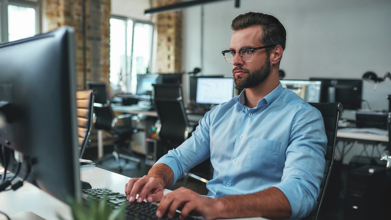 A man with glasses and a blue shirt works on a computer in an office.