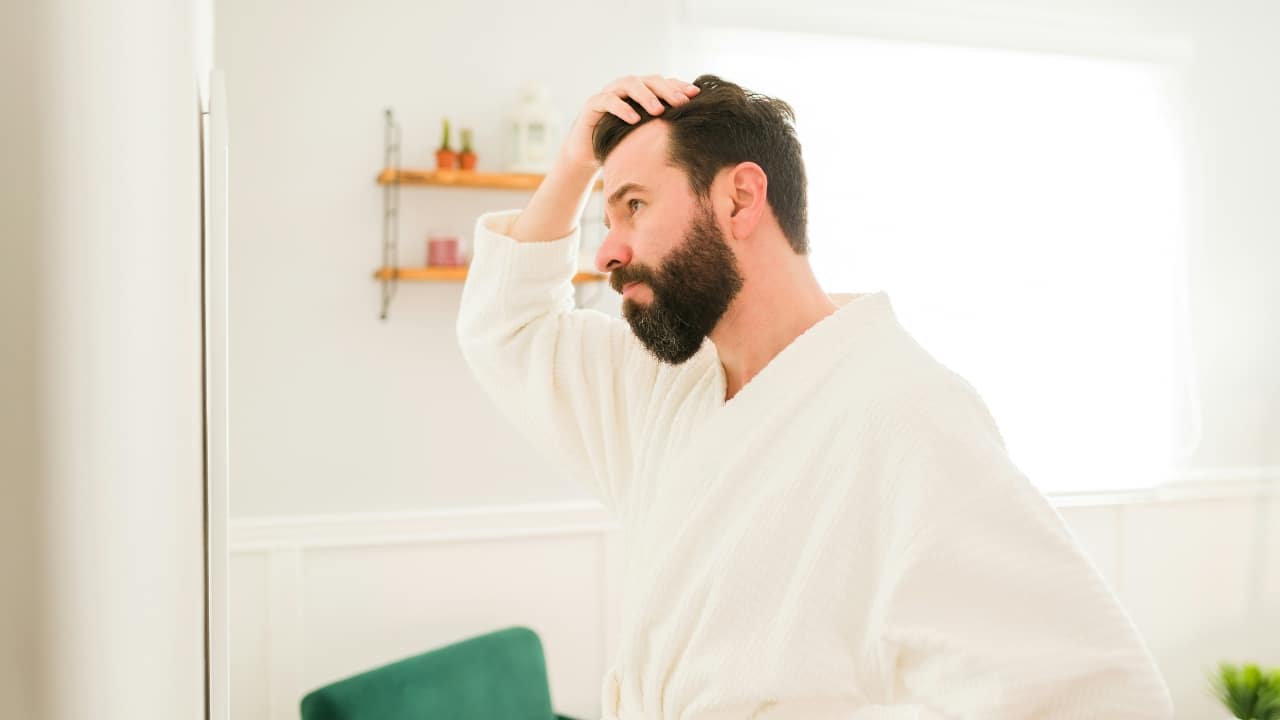 A bearded man in a white bathrobe looks in a mirror and touches his hair.