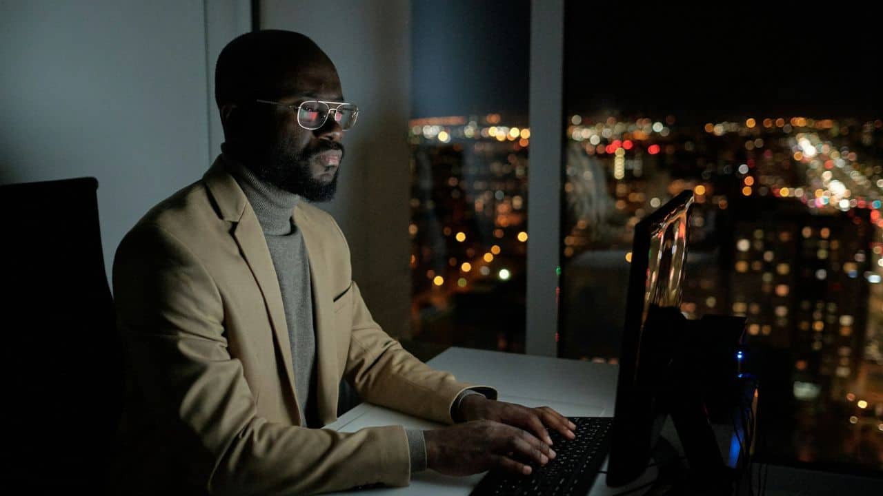 A man in a tan blazer and glasses types on a computer at night.