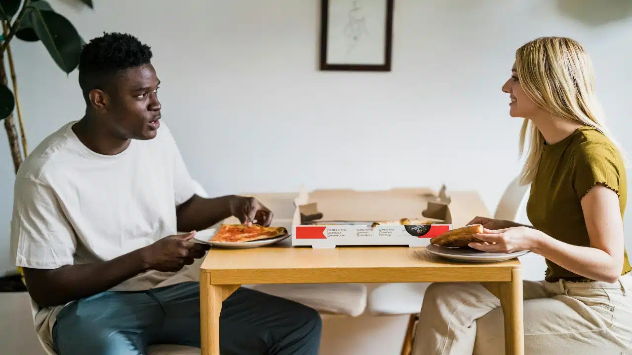 A man and woman sit at a wooden table eating pizza from a cardboard box.