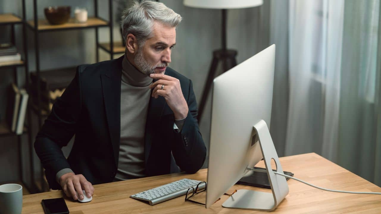 A man in a suit sits at a desk looking at a computer monitor.