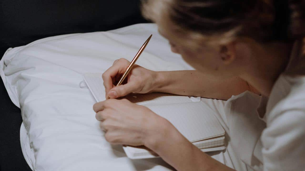 A person writing in a notebook on a bed.