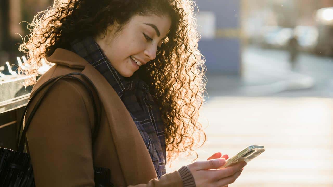 A woman smiling while looking at her phone outdoors.