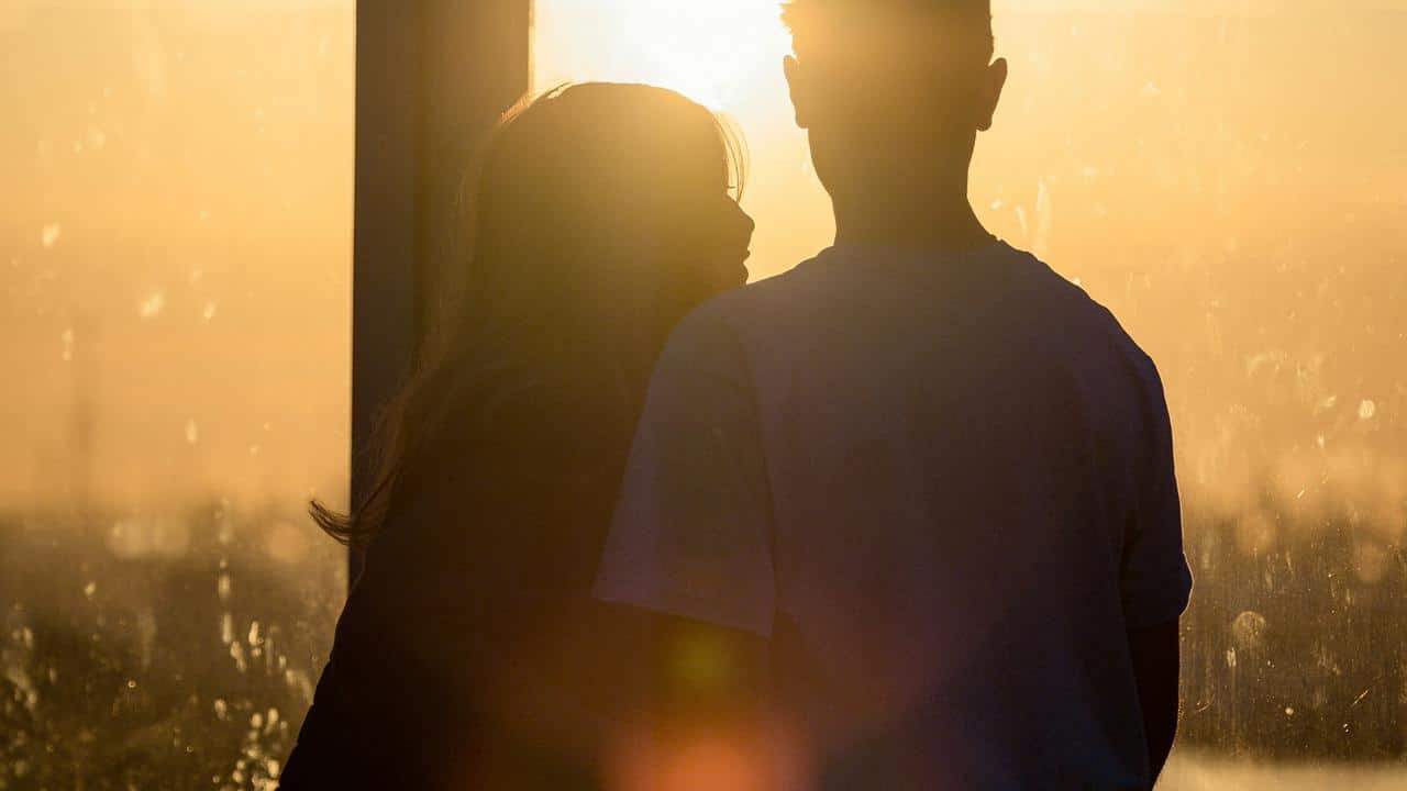 A couple stands close together by a window, silhouetted against a golden sunset.