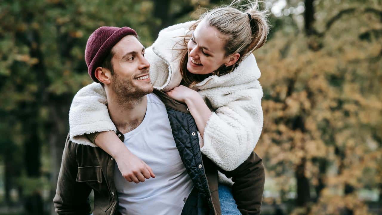 A couple smiling while one gives the other a piggyback ride outdoors.