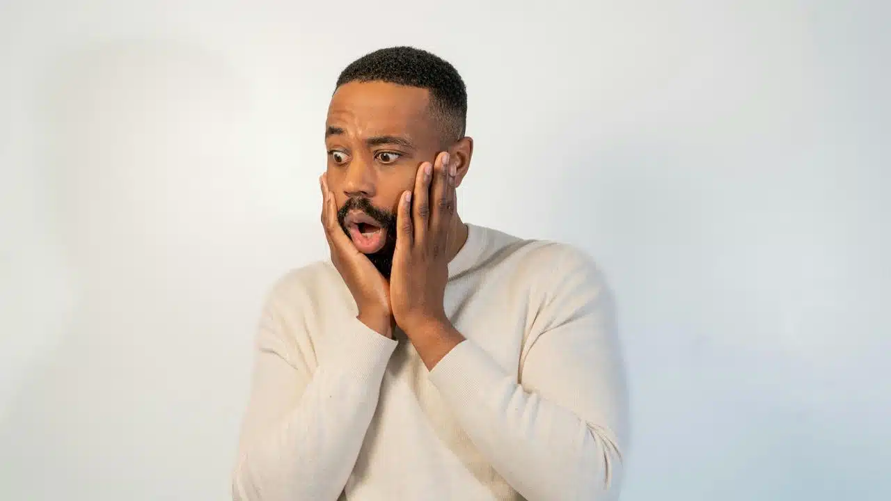 A shocked man holds his face with wide eyes against a plain background.
