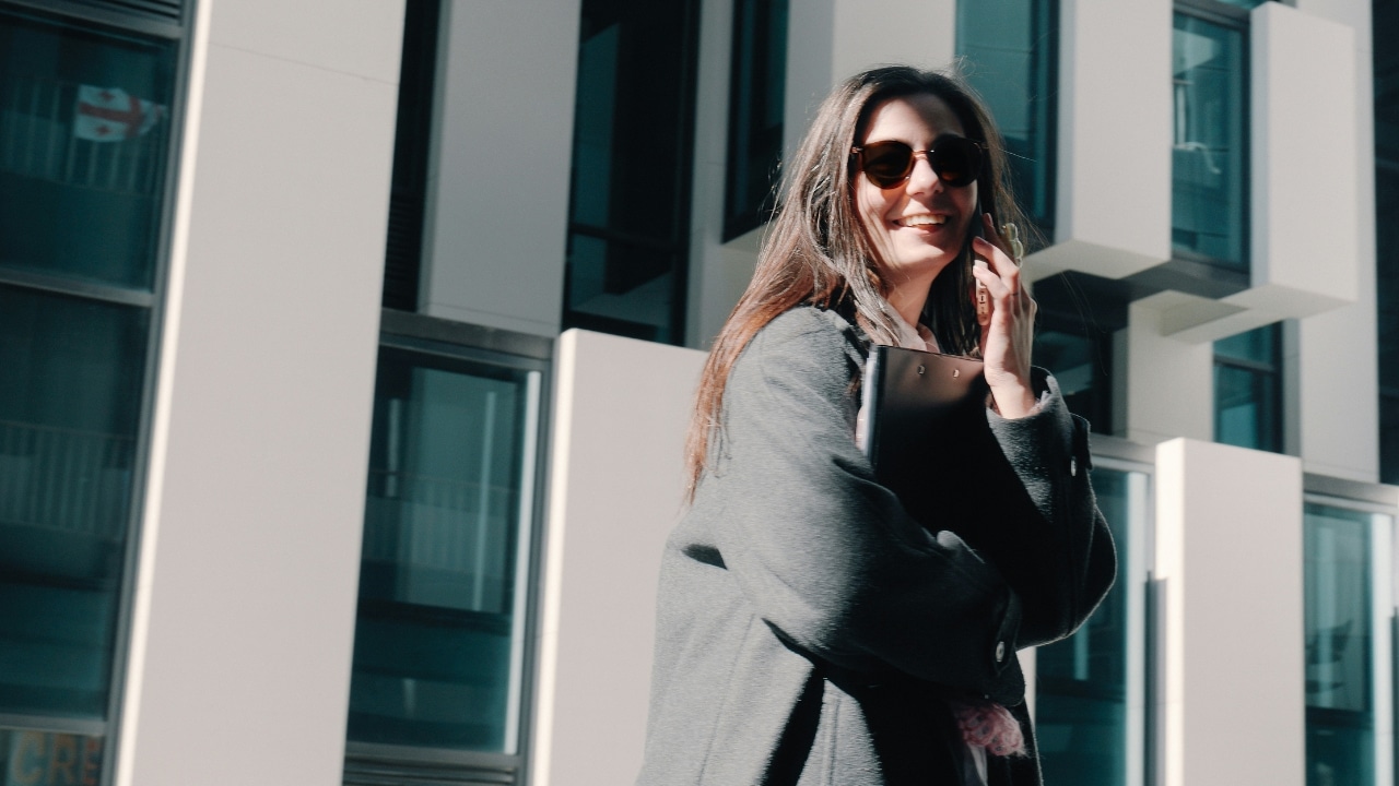 Woman wearing sunglasses and a grey coat walking outside while talking on a smartphone.