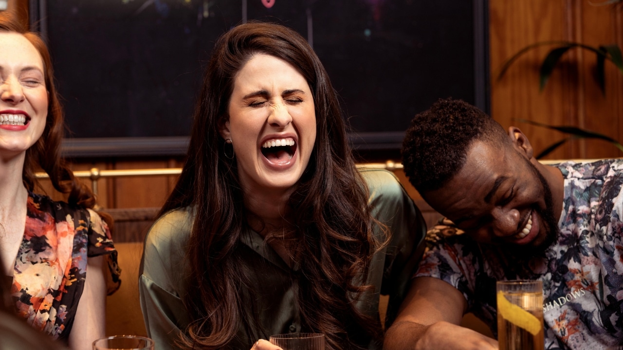 Three people laugh loudly while sitting together at a table in a dimly lit room.