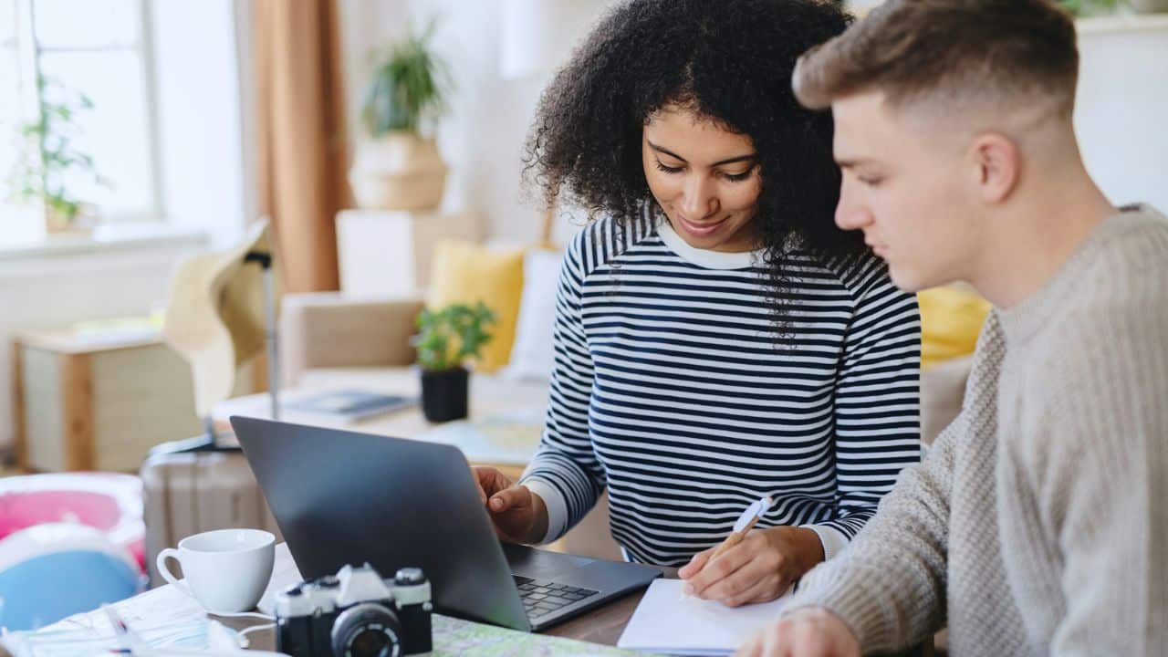 A woman looks at a laptop while a man writes in a notebook nearby.