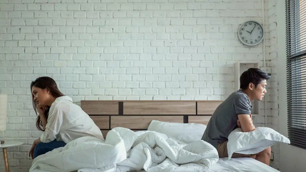 A couple sitting on separate ends of a bed facing away from each other.