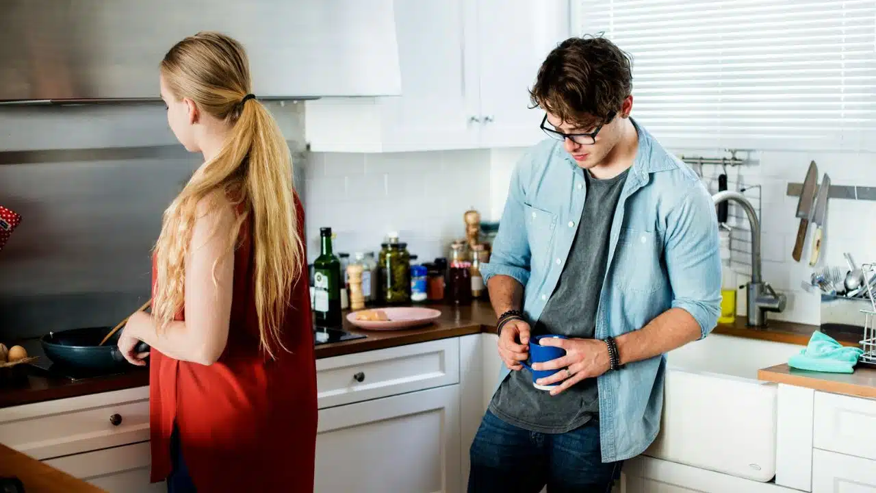A woman cooks at a stove while a man stands nearby holding a blue mug.