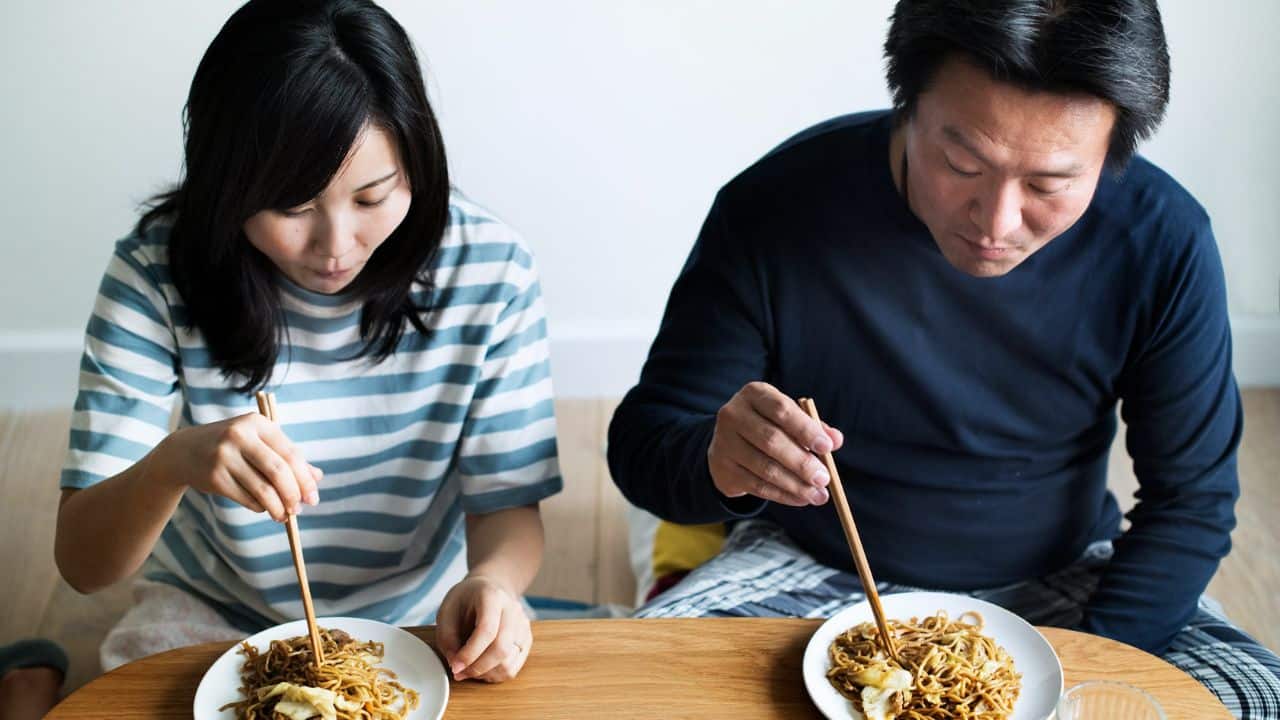 A man and a woman sit at a wooden table eating noodles with wooden chopsticks.
