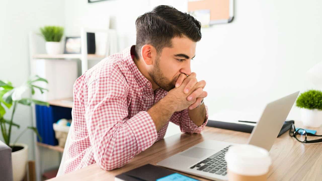 A man in a red and white checkered shirt sits at a desk, looking at a laptop with his hands clasped under his chin.