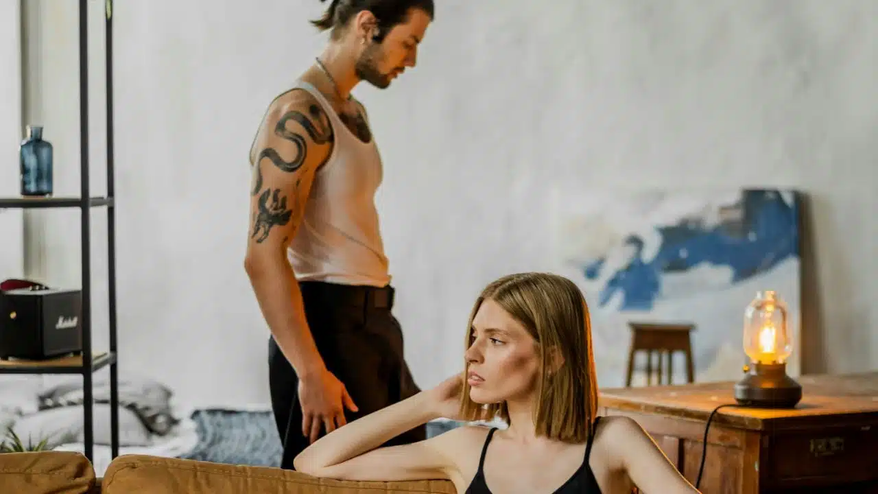 A tattooed man in a tank top stands behind a seated woman looking away.