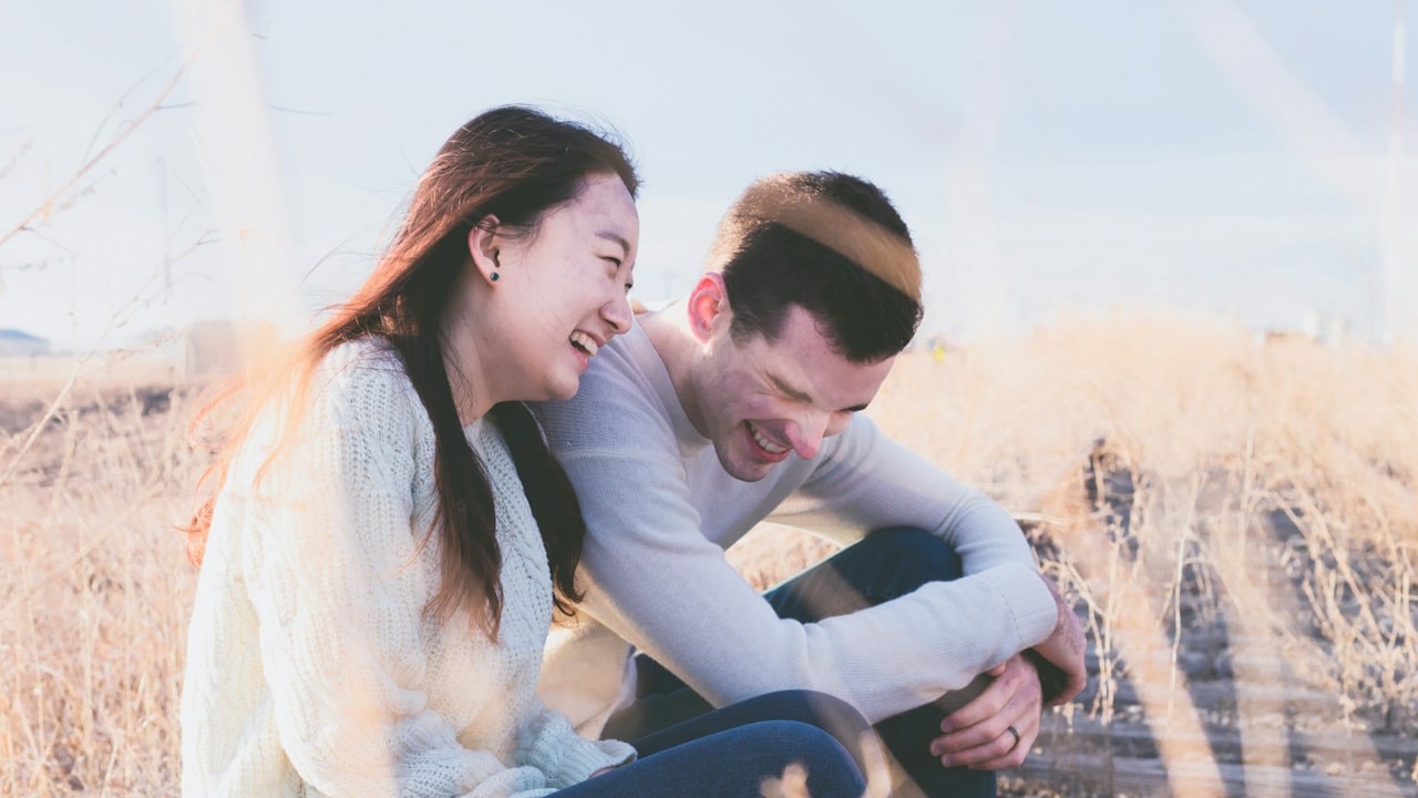 A happy couple sitting close together in a sunny field of dry grass, both laughing joyfully.