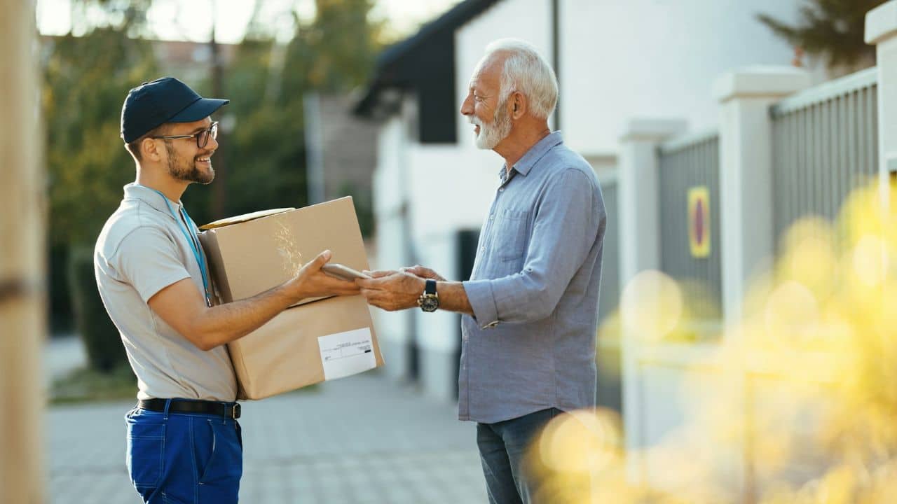 A smiling delivery worker handing two large cardboard boxes to an older man outdoors.