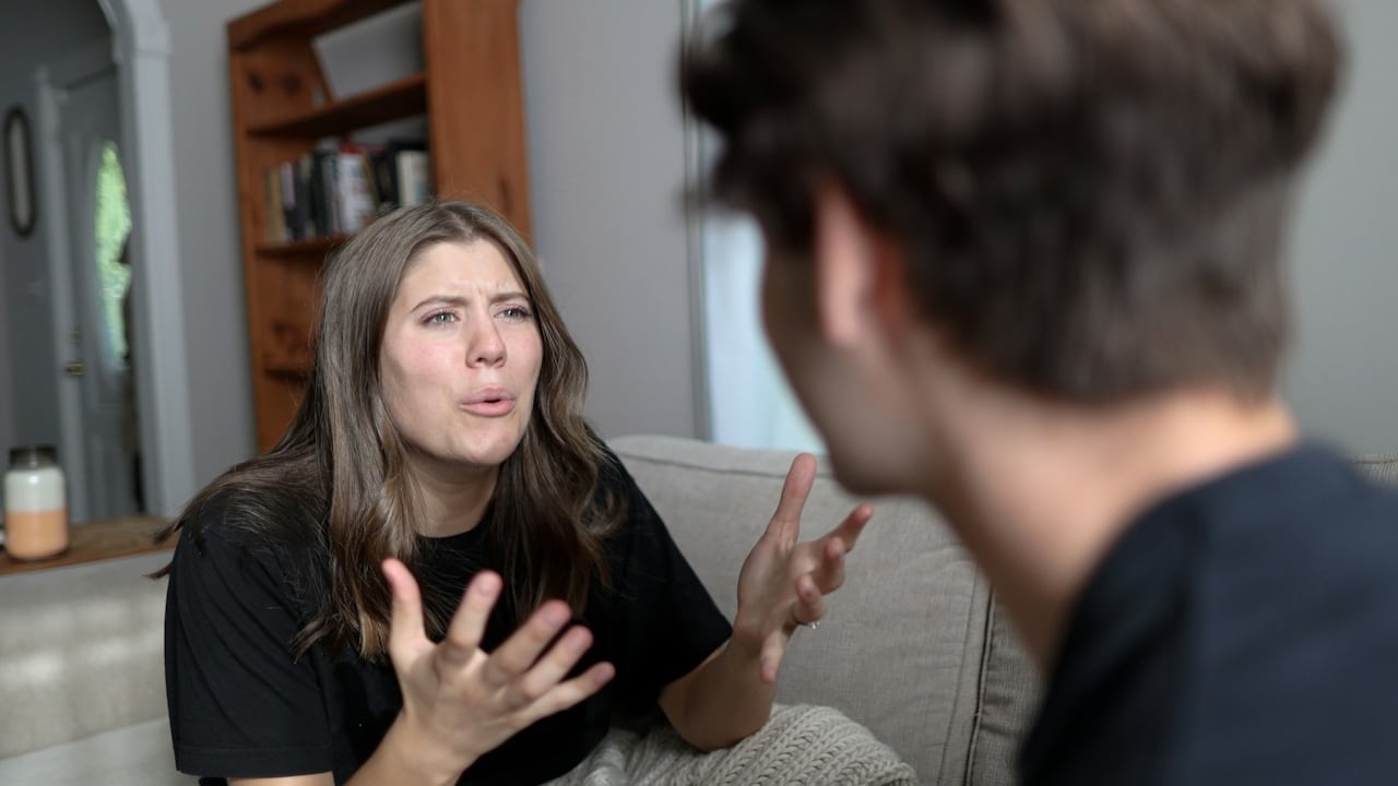A distressed young woman sitting on a couch, wrapped in a blanket, gesticulates while talking to a man